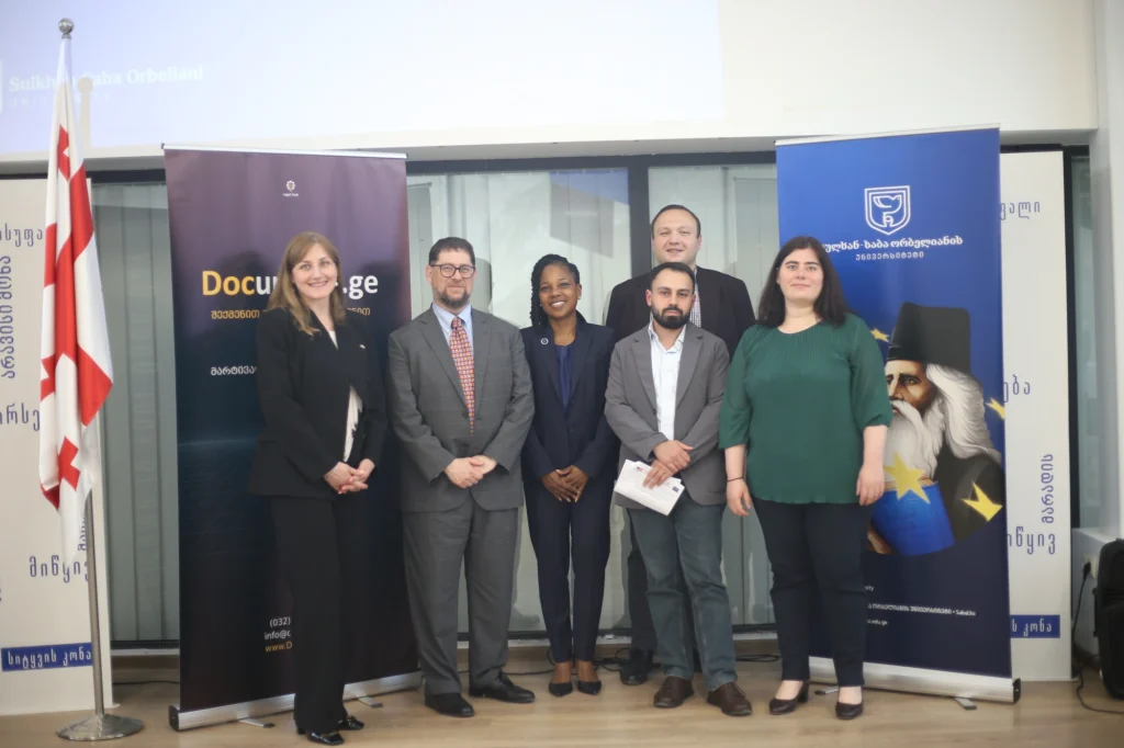 Six participants in the university partnership programme reciprocal visit standing together in front of Document.ge and Sulkhan-Saba Orbeliani University banners.